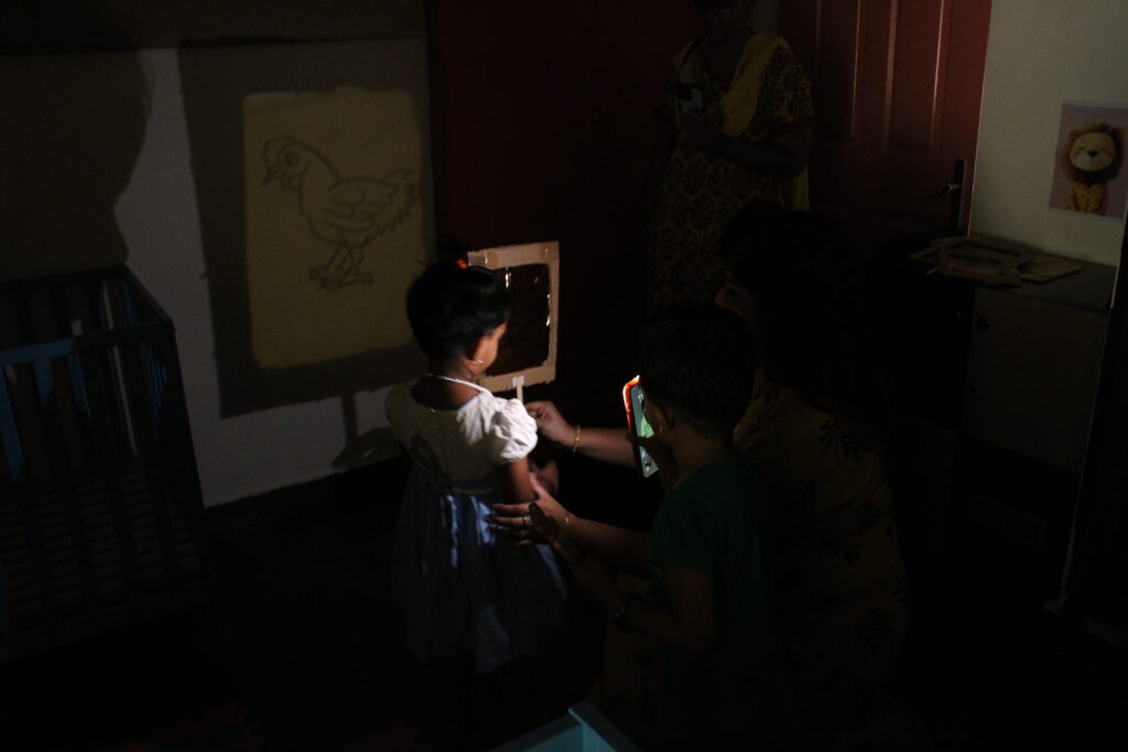 A young child stands in a dimly lit room observing a projected shadow of a chicken drawing on the wall, while adults assist and another child watches with curiosity.