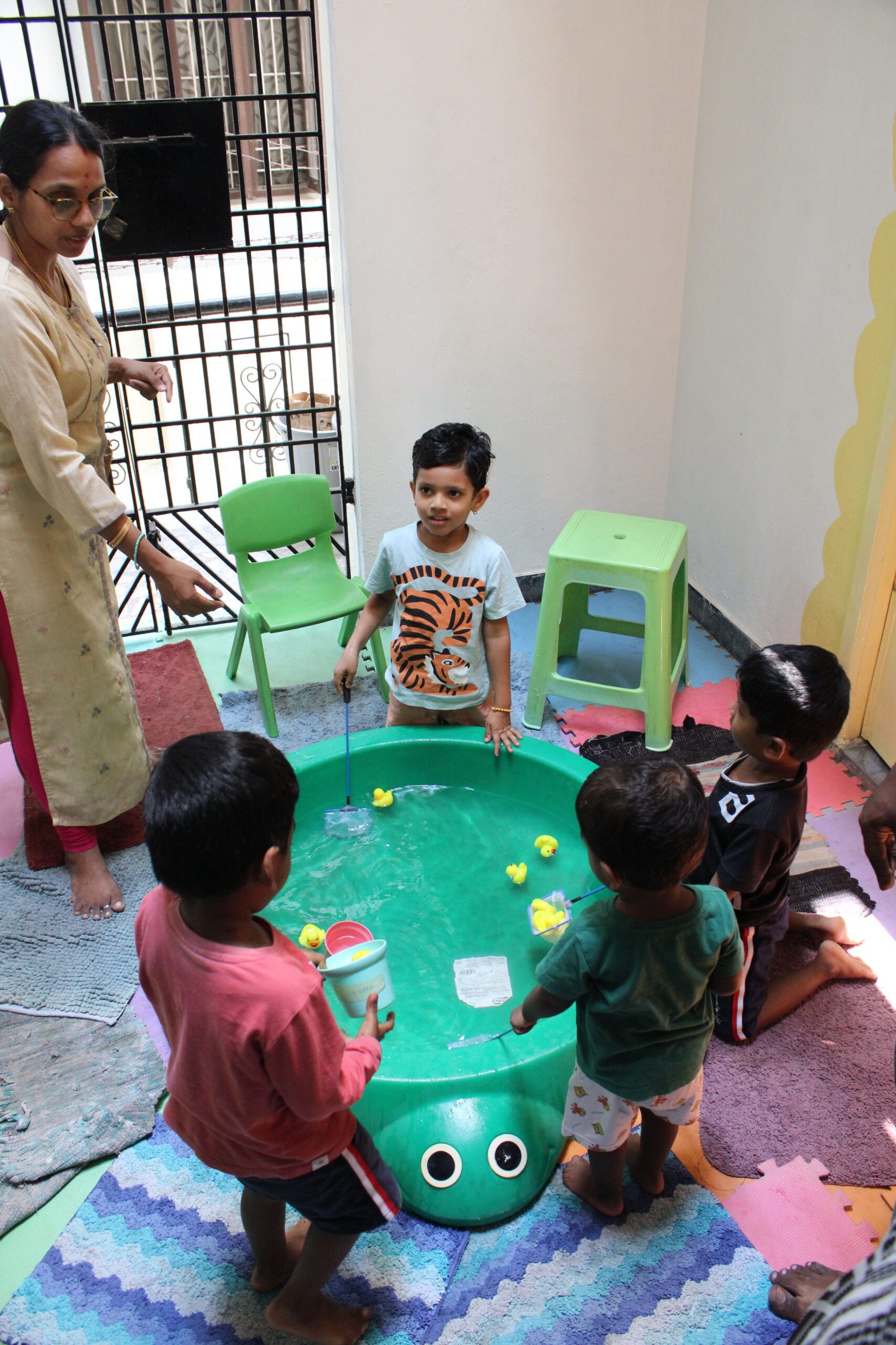 Young children gather around a green water play tub shaped like a turtle, scooping toy ducks under the supervision of a teacher in an indoor play area.
